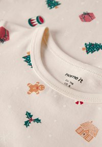 Jasna kremowa koszula z krótkim rękawem z świątecznymi wzorami: zielone choinki, piernikowy ludzik, prezenty, gałązki ostrokrzewu i płatki śniegu. Miękki materiał bawełniany.