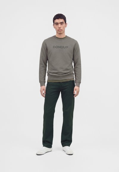 Jeune homme debout portant un sweat-shirt olive avec le texte "DONDUP", pantalon vert foncé et baskets blanches sur un fond clair uni.