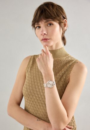 Kobieta z krótkimi brązowymi włosami, ubrana w beżowy, bezrękawnik o fakturze oraz srebrny zegarek na rękę, pozująca z ręką przy brodzie na jednolitym tle.