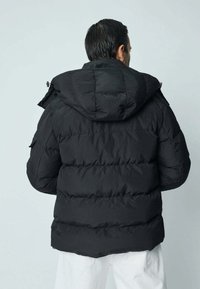 Veste matelassée noire avec capuche, présentant un design matelassé et une texture légèrement brillante. Coutures visibles et sans ornements apparents.