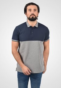Sötét hajú és szakállas férfi, aki sötétkék és szürke rövid ujjú pólót és kék farmert visel, egy sima világos háttér előtt áll.