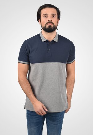 Mand med mørkt hår og skæg iført en navy- og grå kortærmet polo t-shirt samt blå jeans, stående foran en enkel lys baggrund.