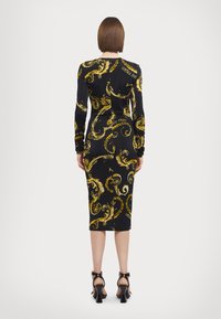 Kroppsnær kjole i svart stoff med dristige gule svirvlende mønstre, lange ermer og kne-lengde kant. Bakvisning viser en strømlinjeformet silhuett og rynker.