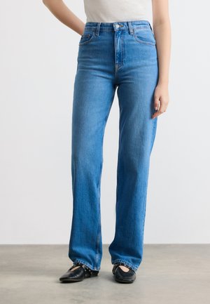 Femme portant un jean bleu taille haute à jambes droites et des ballerines noires cloutées, debout contre un mur blanc uni et un sol gris.