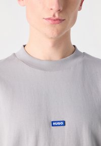 Hellgraues T-Shirt mit Rundhalsausschnitt, ausgestattet mit einem blauen Stoff-Logoetikett mit der Aufschrift "HUGO" in der Mitte der Brust. Glatter Stofftextur.