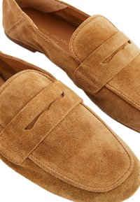 Mocassins en suède beige avec un bout arrondi et une sangle décorative sur le dessus, comportant un détail découpé. Texture douce, couture minimale.