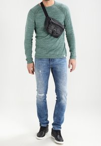 Grünes Langarmshirt, blaue Jeans mit Used-Effekten und schwarze Sneaker. Eine schwarze Umhängetasche, quer über die Schulter getragen. Textur und Details sichtbar.