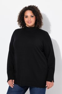 Femme aux cheveux bouclés portant un pull noir à manches longues et un jean bleu, se tenant devant un fond blanc.