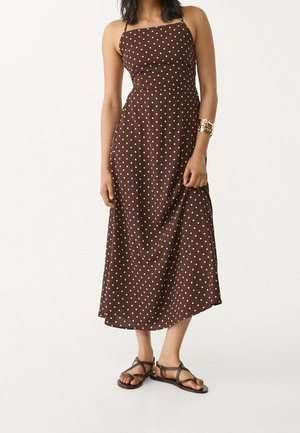 Woman wearing a brown sleeveless maxi dress with white polka dots and black strappy flat sandals, holding dress hem with one hand.