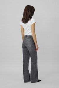 Moteris su pečius siekiančiais rudos spalvos plaukais, vilkinti baltą trumpomis rankovėmis palaidinę, pilkus plačius aukšto liemens džinsus ir juodus batus, atsukta nugara.