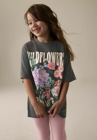 T-shirt gris surdimensionné avec le texte imprimé "Wildflowers" et un design floral coloré. Porté avec des leggings côtelés rose clair. Texture douce.