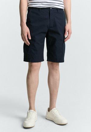 Marinefarbene Cargo-Shorts mit mehreren Taschen, einem Knopfverschluss und Gürtelschlaufen, kombiniert mit weißen Sneakern vor einem neutralen Hintergrund.