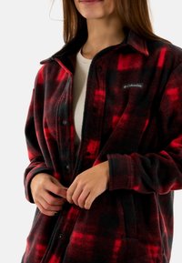 Chaqueta de franela a cuadros rojos y negros con textura suave, botones a presión negros y un parche del logo de Columbia en el lado izquierdo del pecho.