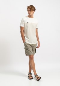 Witte T-shirt met een strandscène-print, gecombineerd met licht khaki cargoshorts. De schoeisel bestaat uit grijze en zwarte verstelbare sandalen.