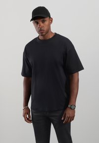 Musta lyhythihainen T-paita pyöreällä pääntiellä, yhdistettynä mustiin housuihin. Asun täydentävät musta lippis ja hopeinen kello, joissa on vähän tekstuuridetaljeja.