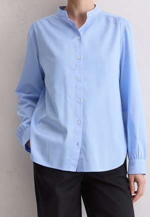 Persona que lleva una blusa de manga larga azul claro con cuello mandarín y botones blancos, combinada con pantalones negros, de pie frente a una pared blanca.