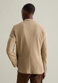 Blazer beige con texture, collo classico, spacco posteriore singolo e tasche laterali, abbinato a pantaloni marroni.