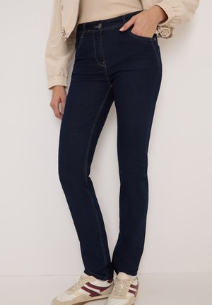 Jean noir foncé à coupe slim, avec une taille haute, des coutures contrastées et des poches avant. Associé à des baskets beiges.