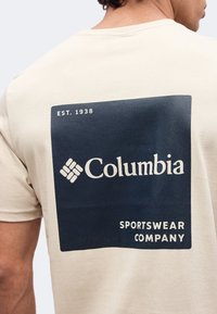 T-shirt beige vu de dos avec un grand carré bleu marine contenant le logo "Columbia Sportswear Company" et le texte "EST. 1938", porté par une personne vue de profil.