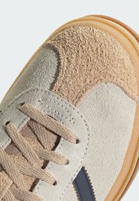 Beige suede sneaker with textured accents, greyish paneling, flat laces, and a rubber sole featuring a light brown edge and navy details.