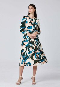 Femme debout portant une robe midi à manches longues avec des motifs floraux abstraits bleus, noirs, blancs et pêche, ainsi que des sandales à talons blancs.