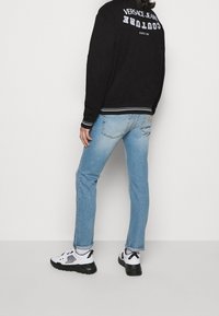 Černá mikina s nápisem „Versace Jeans Couture“ na zádech, v kombinaci se světle modrými džínami a černo-bílými teniskami. Bílé švy jako detaily.