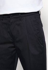 Zwarte broek met een gladde stoftextuur, voorzien van een knoopsluiting, subtiele plooien aan de voorkant en een klassieke rechttoe-rechtaan pasvorm.