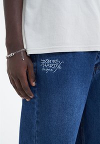 Valkoinen puuvillateeppari sinisten farkkujen päällä, joissa on valkoisella brodeerattu "Don Ed Hardy designs" -logo reidessä ja hopeinen ketjuhandcuff.