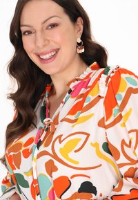 Bluse mit Blumenmuster in leuchtenden Farben – Orange, Pink, Grün – mit rüschigem Kragen und Schleifendetail. Große Creolen ergänzen den Look.