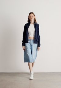 Tmavě modrá zipová bunda s bílými pruhy, bílý crop top a světlé modré volné džíny šortky. Bílé tenisky doplňují outfit.