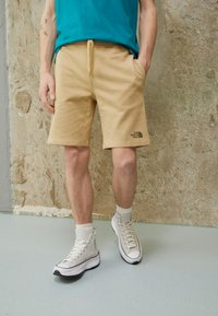 Pantaloni corti beige in cotone con vita elastica e tasche laterali; piccolo logo nero in basso a sinistra. Indossati con sneakers bianche e calzini grigi.