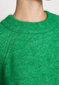 Grön stickad tröja med rund halsringning och strukturerad yta. Tyget ser mjukt ut, med en melerad mönstring och ribbad krage.