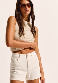 Shorts en denim blanc avec des ourlets roulés, associés à un crop top en tricot beige clair. Le modèle porte des lunettes de soleil sombres. Style simple et décontracté.