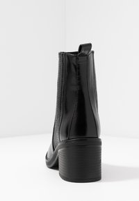 Bota de cuero negro hasta el tobillo con tacón block apilado vista desde atrás sobre una superficie y fondo blancos.
