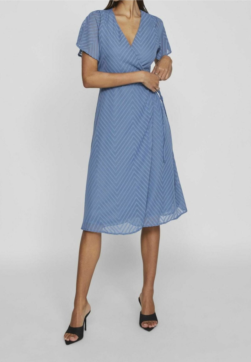 Femme portant une robe portefeuille bleue longueur genou avec manches courtes transparentes et motif chevron, associée à des talons hauts noirs ouverts à l’avant.