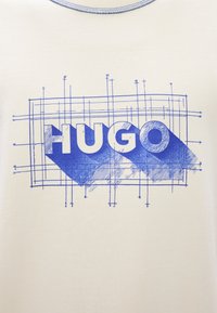 Világos krémszínű póló, amely kék grafikával van díszítve, rajta a "HUGO" szó, rácsvonalakkal és árnyékolási részletekkel strukturálva.