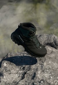 Czarne buty trekkingowe z zielonymi akcentami, wytrzymałą podeszwą, wyściełanym kołnierzem i metalowymi oczkami. Zawieszone nad skalistą powierzchnią z cząstkami kurzu.