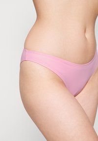 Parte de baixo de biquíni rosa, feita de tecido canelado, apresentando um design de corte baixo e cobertura mínima nas laterais e às costas. Textura suave.