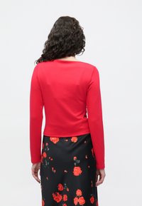 Κόκκινο μπλουζάκι �μακρυμάνικο με στρογγυλή λαιμόκοψη, λείο ύφασμα, συνδυασμένο με μαύρη φούστα που έχει λουλουδάτο σχέδιο σε κόκκινο και πράσινο.