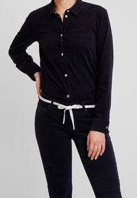 Persona che indossa una camicia nera con bottoni e pantaloni neri, con una cintura bianca legata in vita, su uno sfondo grigio uniforme.