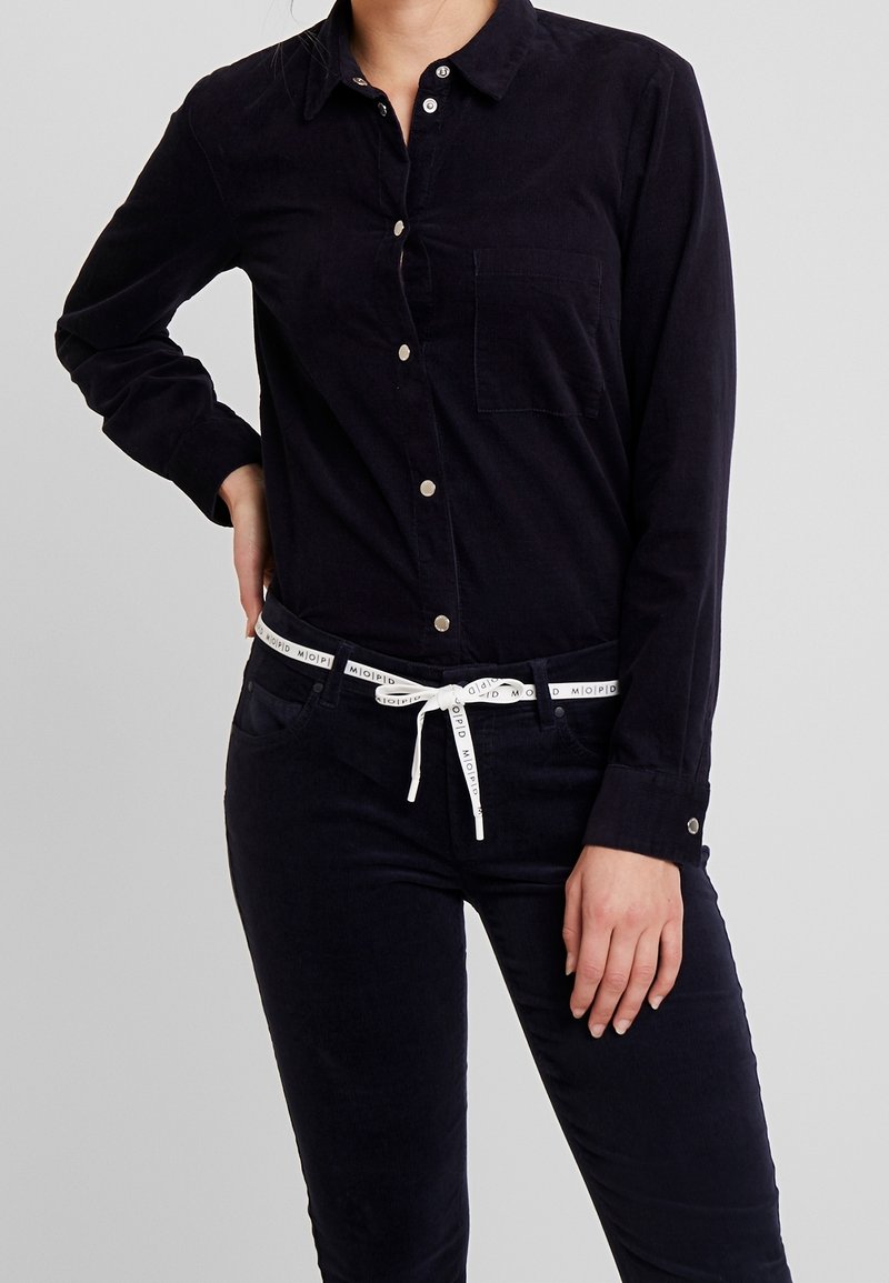 Persona che indossa una camicia nera con bottoni e pantaloni neri, con una cintura bianca legata in vita, su uno sfondo grigio uniforme.