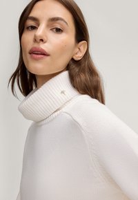 Weißer gerippter Rollkragenpullover mit einem strukturierten Design und einem kleinen goldenen Akzent am Kragen. Weicher Strickstoff, schlanker Schnitt.