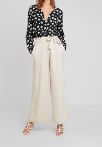 Blouse noire à imprimé marguerites blanches, col en V, manches longues, associée à un pantalon beige à jambes larges avec ceinture et poches. Sandales blanches à lanières.