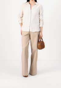 Light beige linen button-up shirt with two chest pockets, paired with light beige wide-leg trousers. Holding a small brown textured handbag.