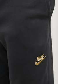 Black sweatpants with a soft texture, featuring a gold embroidered Nike logo on the left thigh. Simple design, standard fit.