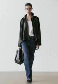 Frau, die geht und eine schwarze Lederjacke, ein weißes Tanktop, blaue Jeans, schwarze Schuhe trägt und eine lässige schwarze Ledertasche hält.