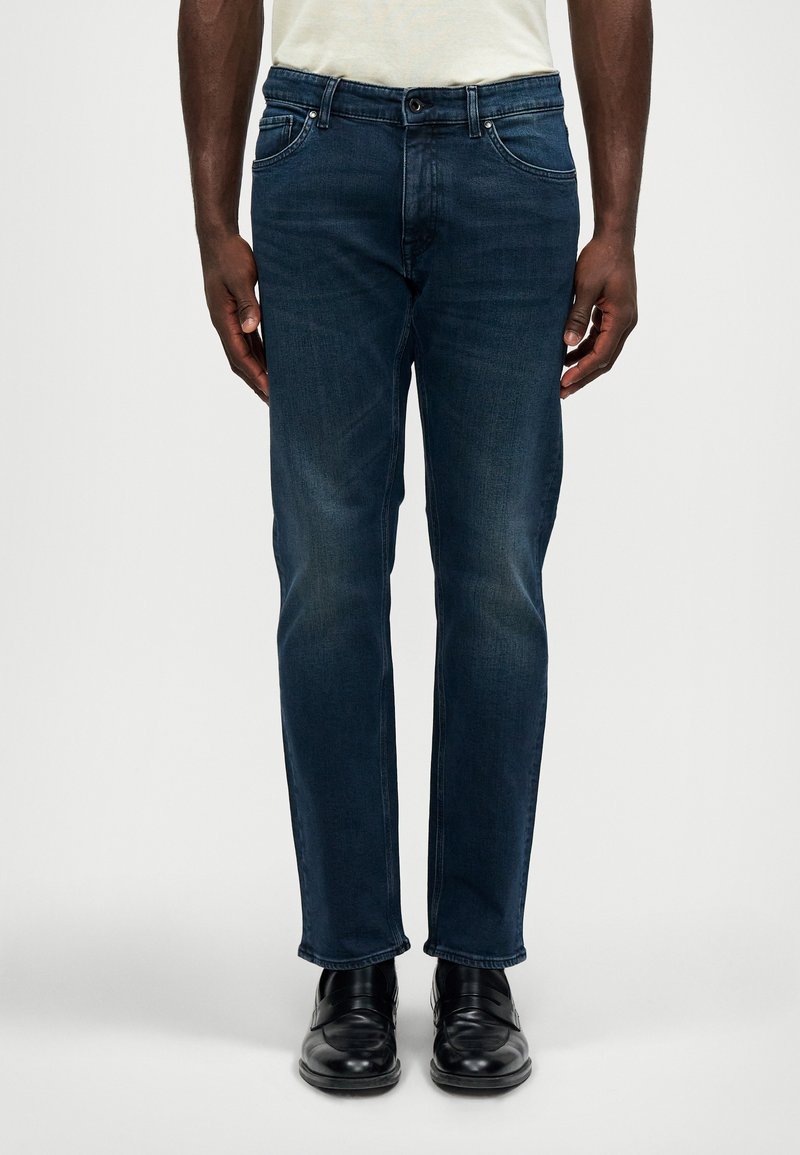 Homem vestindo jeans escuros slim fit, camisa branca e sapatos de couro pretos, de pé contra um fundo claro liso.