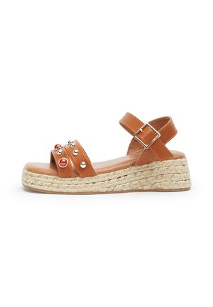 Sandalo con zeppa in pelle marrone con fibbia alla caviglia, decorazioni con borchie e perline sulle fasce frontali e suola piattaforma in juta intrecciata.