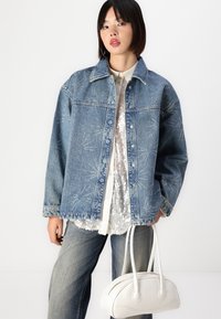 Denim jakna s srebrnim vezenjem z zvezdnim vzorcem, svetlo modre barve, v kombinaciji s srajco s bleščicami in svetlo modrimi kavbojkami. Bele ukrivljene torbice držite v roki.