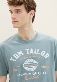 Gaiši zils kokvilnas T-krekls ar apaļu kaklu. Iezīmēts ar "TOM TAILOR" logotipu baltā un oranžā krāsā apļveida emblēmas dizainā. Īsas piedurknes.